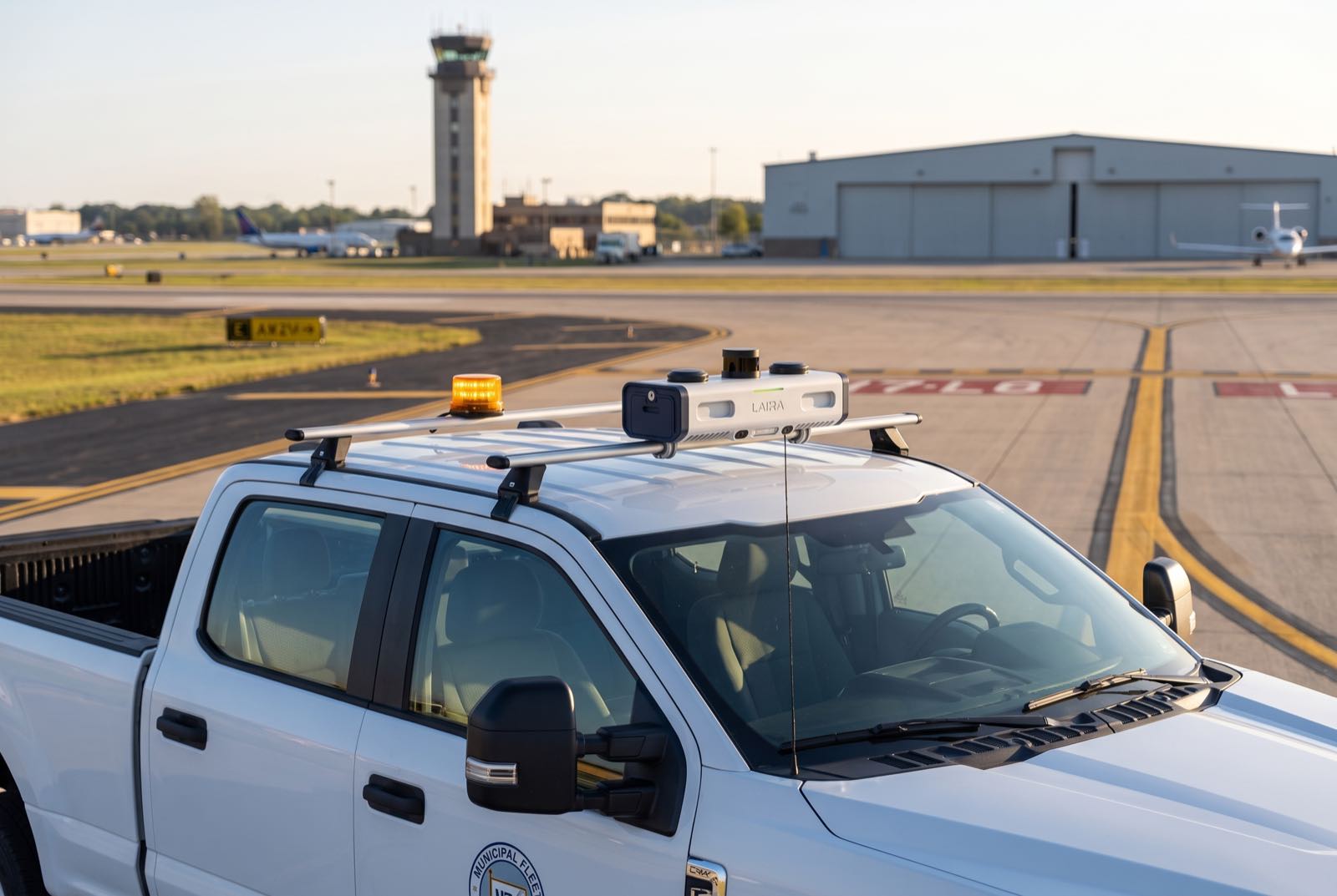 An airfield-operations pickup carrying Airwai's LAIRA sensor rig on an active taxiway at golden hour, control tower behind.